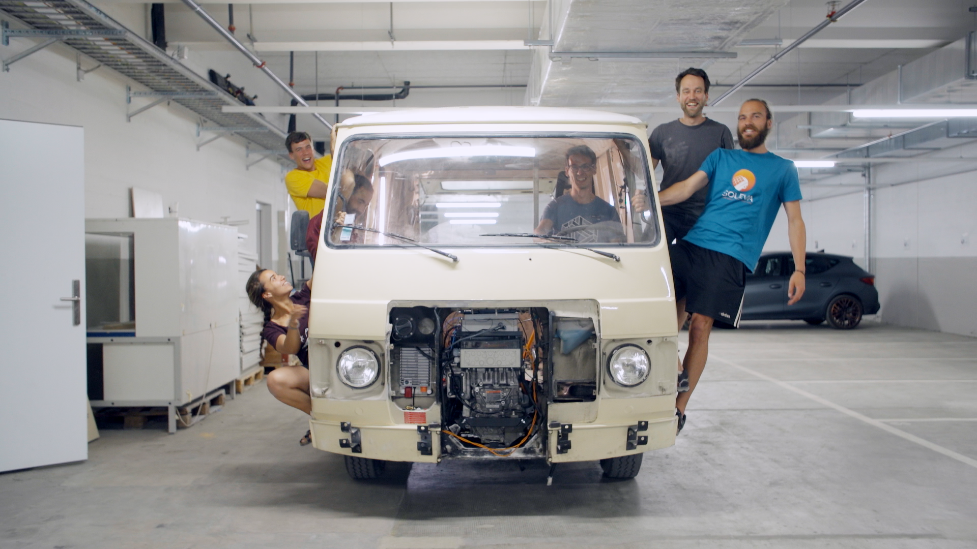 Un van de face sans calandre avec moteur électrique visible et des personnes qui se tiennent sur les côtés